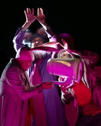 Vergrößerte Ansicht von Drei Personen in bunter Kleidung, eine hält eine weiße Teekanne, eine andere formt mit den Händen ein Dreieck über dem Kopf.