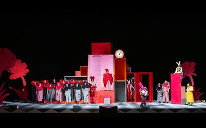 Vergrößerte Ansicht von Bühnenbild mit bunten Kuben, Uhr, mehreren Darstellern in fantasievollen Kostümen, darunter ein Hase und Figuren in Rot und Gelb.