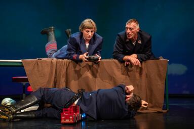 Vergrößerte Ansicht von Ein Feuerwehrmann liegt vor einem Tisch und schlägt die Hände über dem Kopf zusammen. Zwei Mitarbeitende der Feuerwehr schauen auf ihn vom Tisch herrab.