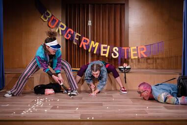 Vergrößerte Ansicht von Man sieht drei Personen in bunter Kleidung. Die Person links steht breitbeinig da und hält eine Blockflöte wenige Meter über dem Boden. In der Mitte steht eine weitere Person, die die Flöte wenige Zentimeter vor dem Boden hält vor Popcorn. Auf der…