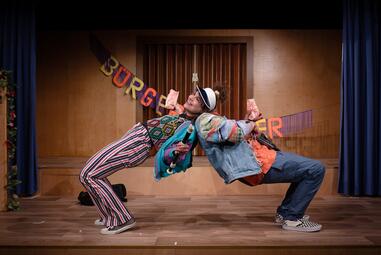 Vergrößerte Ansicht von Zwei Personen in bunter Kleidung lehnen ihre Oberkörper nach hinten und stützen sich gegenseitig mit ihren Oberkörpern.Die Person links hält eine kleine Tüte Popcorn in einer Hand und eine Blockflöte in der anderen Hand fest. Sie lächelt. Die andere…