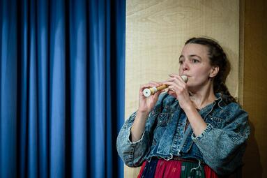 Vergrößerte Ansicht von Eine Frau mit braunen Haaren und einer Jeansjacke lehnt sich an eine Wand an und spielt dabei Blockflöte. Sie wirkt sehr konzentriert. Im Hintergrund ist ein blauer Vorhang.