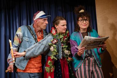 Vergrößerte Ansicht von Drei Personen stehen nebeneinander vor einem dunkelblauen Vorhang. Die Person, die links steht, hält eine Zeitung in der Hand.Die anderen beiden halten eine Blockflöte in der Hand und schauen kritisch in die Zeitung.