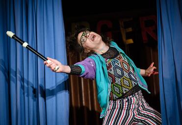 Vergrößerte Ansicht von Eine Frau mit dunkelbraunen Haaren, bunter Kleidung und einer Brille streckt ihren Arm aus. In der Hand hält sie eine Blockflöte wie einen Zauberstab. Sie scheint etwas zu sagen.