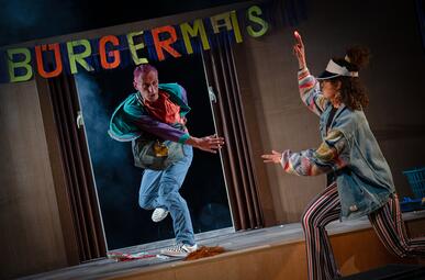 Vergrößerte Ansicht von Ein Mann in bunter Kleidung springt auf einer Bühne. Auf der rechten Seite steht eine Frau, die einen Finger zur Decke streckt und seitlich zur Kamera steht.