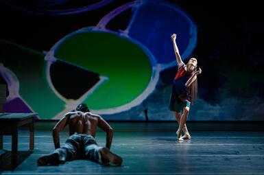 Vergrößerte Ansicht von Auf der linken Seit liegt eine Person mit dem Rücken zum Publikum. Die Person liegt bäuchlings und trägt eine schwarze Hose und kein T-Shirt. Sie hat die Unterarme auf den Boden gestützt. Neben ihr steht eine Person mit überkreuzten Beinen und…