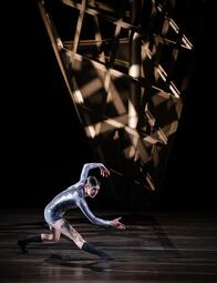 Vergrößerte Ansicht von Es ist eine Person in einem silbernen Jumpsuit zu sehen, die seitlich zum Publikum steht und die Beine beugt. Einen Arm streckt sie nach vorne, den anderen hält sie über ihren Kopf.