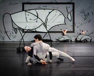 Vergrößerte Ansicht von Eine Person sitzt mit dem Rücken zur Kamera und in einem mehrfarbigen Ganzkörperanzug. Auf dieser Person lehnt eine andere Person, die sich mit einer Hand an ihr festhält und mit der anderen Hand abstützt. Im Hintergrund sind zwei Tänzerinnen zu…