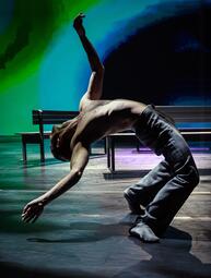 Vergrößerte Ansicht von Eine Person in grauer Hose und ohne Oberteil lehnt mit gebeugten Beinen den gesamten Oberkörper nach hinten. Eine Hand berührt beinahe den Boden und den anderen Arm streckt die Person zur Decke.Im Hintergrund sind weiße Bänke zu sehen.