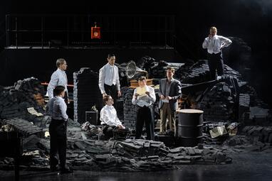 Vergrößerte Ansicht von Sieben Männer in verschiedenen Arten von Abendkleidung stehen in den Ruinen von Mauern. Alle halten den Blick auf den siebten Mann gerichtet, der ein Stück Papier in der Hand hält und davon vorliest.