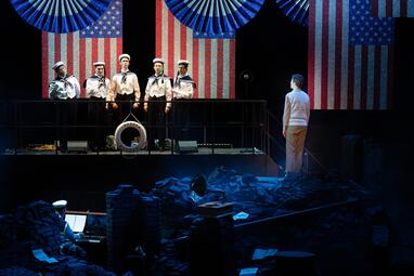 Vergrößerte Ansicht von Fünf Personen in Matrosenuniformen stehe vor zwei von der Decke herabhängenden Flaggen der USA. Sie scheinen auf einem Boot zu sein, was am Rettungsring vor ihnen gezeigt wird. Sie werden beobachtet von einer Person mit dem Rücken zum Publikum, in…