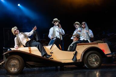 Vergrößerte Ansicht von Von fünf Männern auf dem Bild, alle in Fracks ohne Jacken, mit Kopfbedeckungen und Sonnenbrillen, sitzt einer am Steuer. Ein anderer lehnt sich auf der Motorhaube nach hinten, während die anderen drei hinter dem Auto stehen und wie alle anderen in…