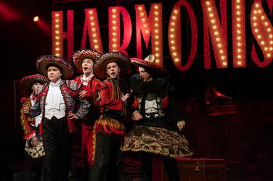 Vergrößerte Ansicht von Fünf Männer in traditioneller, spanischer Kleidung mit Sombreros auf den Köpfen drängen sich am linken Bildrand zusammen. DIe rote Farbe ihrer Kleidung spiegelt sich in der roten Farbe des Schriftzugs &quot;Harmonists&quot; hinter ihnen and er Bühnenwand.