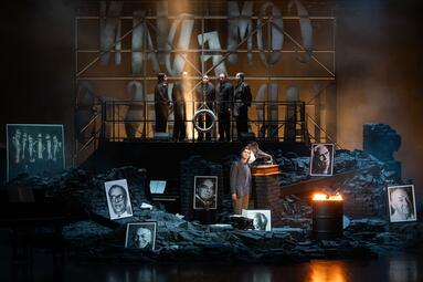 Vergrößerte Ansicht von Die sechs Sänger stehen in den abgebrandten Ruinen von Häusern. Fünf von ihnen stehen im Hintergrund auf einem Gerüst vor den kaputten Wörtern &quot;Comedian Harmonists&quot;, der sechste Mann unten in den Ruinen vor einer brennenden Tonne. Die gesamte…