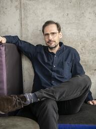 Vergrößerte Ansicht von Portrait von Jochen Schliemann auf einer Bank sitzend vor einer grauen Betonwand. Mit dunkelgrünem Hemd, Jeans und Brille, Bart und dunklen kurzen Haaren.