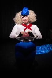 Vergrößerte Ansicht von Dragqueen Miss Sara Jevo trägt eine blaue Mütze, weiße Bluse mit rotem Halstuch und blauen Rock, hält einen roten Reisepass.