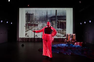 Vergrößerte Ansicht von Dragqueen Miss Sara Jevo steht auf einer Bühne, trägt ein rotes Outfit mit weitem Hosenrock und hält einen langen Stock in der rechten Hand. Im Hintergrund ist eine großflächige Projektion einer Stadtlandschaft zu sehen.