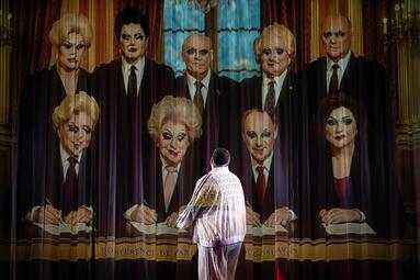 Vergrößerte Ansicht von Dragqueen Miss Sara Jevo steht vor einer großen Leinwand mit einem historischen Gruppenfoto von Männern in Anzügen und Krawatten, die an einem Tisch sitzen.