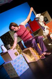 Vergrößerte Ansicht von Person in gestreiftem T-Shirt und gestreifter Hose sitzt auf buntem Würfel und hält zwei kleine weiße Ventilatoren in der Hand.