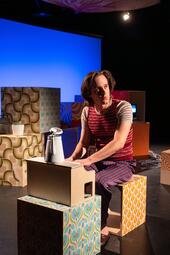 Vergrößerte Ansicht von Person sitzt auf einem gemusterten Würfel, trägt gestreiftes T-Shirt und karierte Hose, vor ihr steht eine weiße Kaffeekanne auf einem Karton.