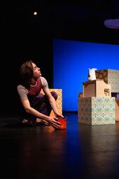 Vergrößerte Ansicht von Person in gestreiftem T-Shirt und Hose hockt auf Bühne und hält roten Würfel, daneben bunte Würfel mit Blumenmuster und weiße Kanne.