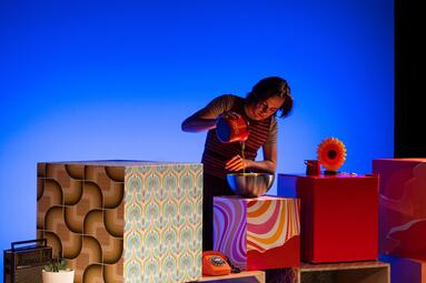 Vergrößerte Ansicht von Person gießt Flüssigkeit aus einem roten Kännchen in eine Metallschüssel, umgeben von bunten, gemusterten Würfeln und einem orangefarbenen Telefon auf blauem Hintergrund.