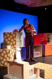 Vergrößerte Ansicht von Person in gestreiftem Oberteil und gestreifter Hose steht auf einer Bühne mit bunten, gemusterten Würfeln und Requisiten.