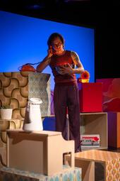 Vergrößerte Ansicht von Person mit rot-weiß gestreiftem Oberteil und rot-lila gestreifter Hose hält roten Telefonhörer ans Ohr, steht vor bunten, gemusterten Würfeln und Möbeln.