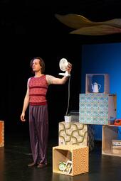 Vergrößerte Ansicht von Person in gestreiftem rotem Oberteil und gestreifter Hose hält einen kleinen weißen Ventilator in der linken Hand, steht neben bunten Würfeln mit Mustern und Haushaltsgegenständen.