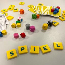 Vergrößerte Ansicht von Würfel, Spielsteine und Spielkarten auf einer grauen Oberfläche. Fünf gelbe Spielplättchen bilden das Wort "Spiel".