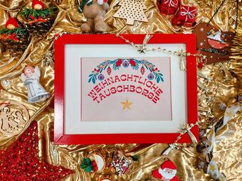 Vergrößerte Ansicht von In einem roten Bilderrahmen steht: Weihnachtliche Tauschbörse. Der Rahmen  liegt auf goldfarbenem Stoff inmitten von Weihnachtsdeko.