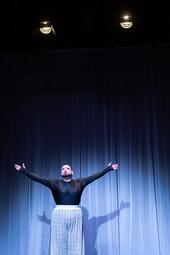 Vergrößerte Ansicht von Miss Sara Jevo steht mit ausgebreiteten Armen vor einem blauen Bühnenvorhang, trägt ein schwarzes Oberteil und eine weiße Hose.