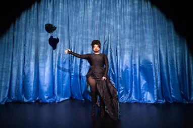 Vergrößerte Ansicht von Miss Sara Jevo in einem schwarzen, asymmetrischen Kleid mit hohem Schlitz steht vor einem blauen Vorhang und wirft einen schwarzen Hut mit grünen Akzenten in die Luft.