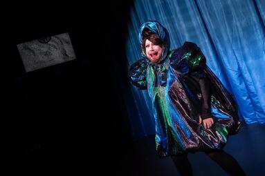 Vergrößerte Ansicht von Miss Sara Jevo in schimmerndem, mehrfarbigem Kleid mit Puffärmeln und passendem Kopftuch vor blauem Vorhang auf dunkler Bühne.