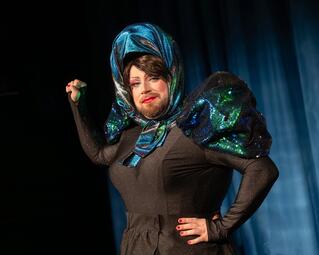 Vergrößerte Ansicht von Miss Sara Jevo trägt ein schwarzes, eng anliegendes Kleid mit großen, glitzernden, blau-grünen Puffärmeln und ein passendes Kopftuch und hält die rechte Faust in die Luft.