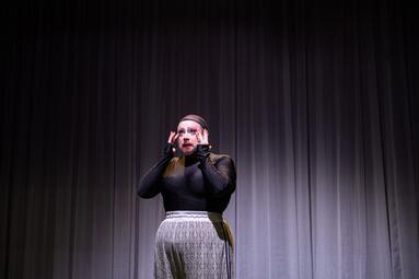 Vergrößerte Ansicht von Miss Sara Jevo steht auf einer Bühne vor einem grauen Vorhang, trägt ein schwarzes Oberteil und einen hellen, gemusterte Hose.