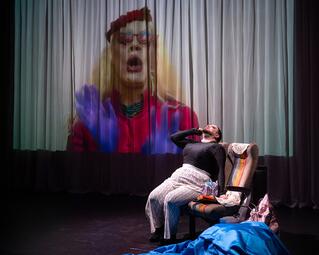 Vergrößerte Ansicht von Miss Sara Jevo sitzt auf einem bunten Stuhl, hält sich die Hand an den Kopf, vor einem Vorhang mit einer großen Projektion einer Person in rotem Oberteil und blauen Handschuhen.