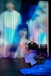 Vergrößerte Ansicht von Miss Sara Jevo sitzt auf einem Stuhl mit buntem Überwurf vor einer Leinwand, auf der verschwommene, farbige Figuren projiziert sind.