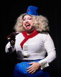 Vergrößerte Ansicht von Miss Sara Jevo mit lockiger blonder Perücke, blauem Hut mit rotem Stern, rotem Halstuch, weißer Bluse und blauer Hose mit Strasssteinen hält einen Pass in der Hand.