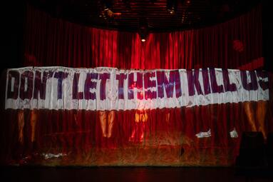 Vergrößerte Ansicht von Roter Bühnenvorhang mit einer Projektion auf der ein großes weißes Banner abgebildet ist, auf dem in schwarzen und roten Buchstaben steht: &#039;DON&#039;T LET THEM KILL US&#039;.