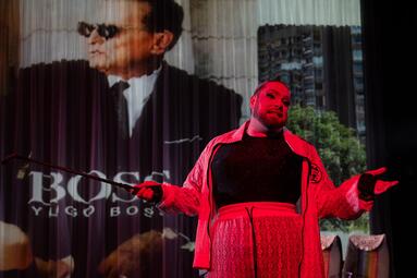 Vergrößerte Ansicht von Miss Sara Jevo steht in rötlichem Licht vor einer Projektion mit einem Mann im Anzug und dem Schriftzug &#039;BOSS YUGO BOSS&#039;.