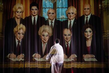 Vergrößerte Ansicht von Miss Sara Jevo steht vor einer Projektion mit einer historischen Gruppenaufnahme von Personen in Anzügen, die an einem Tisch sitzen.