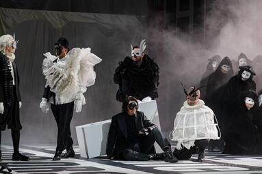 Vergrößerte Ansicht von Mehrere Personen in schwarz-weißer, avantgardistischer Kleidung auf einer Bühne mit Nebel, darunter ein sitzender Mann in schwarzem Anzug und eine Person in weißem, gerüstartigem Kostüm mit Hörnern.