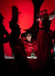 Vergrößerte Ansicht von Mehrere Personen in dunkler Kleidung und Kopfbedeckungen vor rotem Hintergrund, eine Person mit Hut auf dem Kopf, auf dem Tisch steht eine grüne Glasflasche.