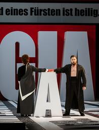 Vergrößerte Ansicht von Zwei Personen in schwarzer Kleidung berühren sich mit einer Hand auf einem großen weißen Buchstaben A auf einer Bühne mit großem rotem und weißem Hintergrund.