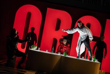 Vergrößerte Ansicht von Drei Personen in Kostümen an einem Tisch mit Flaschen, vor rotem Hintergrund mit großen Buchstaben &#039;ORGI&#039;.