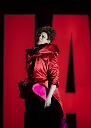 Vergrößerte Ansicht von Person in rotem, glänzendem, voluminösem Oberteil mit großen Falten und schwarzem Rock vor rotem Hintergrund mit schwarzem Schriftzug.