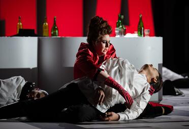 Vergrößerte Ansicht von Eine Person in rotem Kleid und roten Handschuhen kniet auf dem Boden und hält eine andere Person in weißem Hemd und schwarzer Hose, die reglos auf dem Boden liegt. Im Hintergrund steht eine weiße Theke mit mehreren grünen und braunen Flaschen davor…