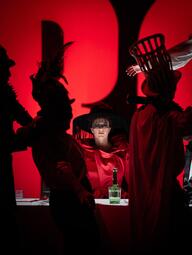 Vergrößerte Ansicht von Im Vordergrund sind mehrere Personen in dunkler Kleidung mit auffälligen Kopfbedeckungen zu sehen, darunter eine mit einem hohen, gefiederten Hut und eine andere mit einem korbartigen Hut auf dem Kopf. Im Hintergrund sitzt eine Person in rotem Kleid…