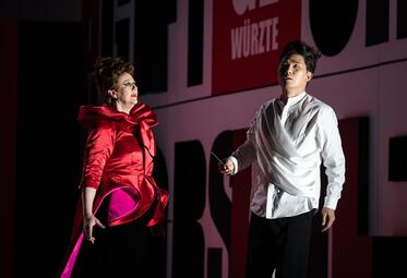 Vergrößerte Ansicht von Eine Frau trägt ein auffälliges rotes Kleid mit großen, voluminösen Schleifen- und Rüschenapplikationen an den Schultern und Hüften. Ein Mann steht neben ihr in einem weißen, locker sitzenden Hemd mit asymmetrischem Schnitt und schwarzen Hosen.…